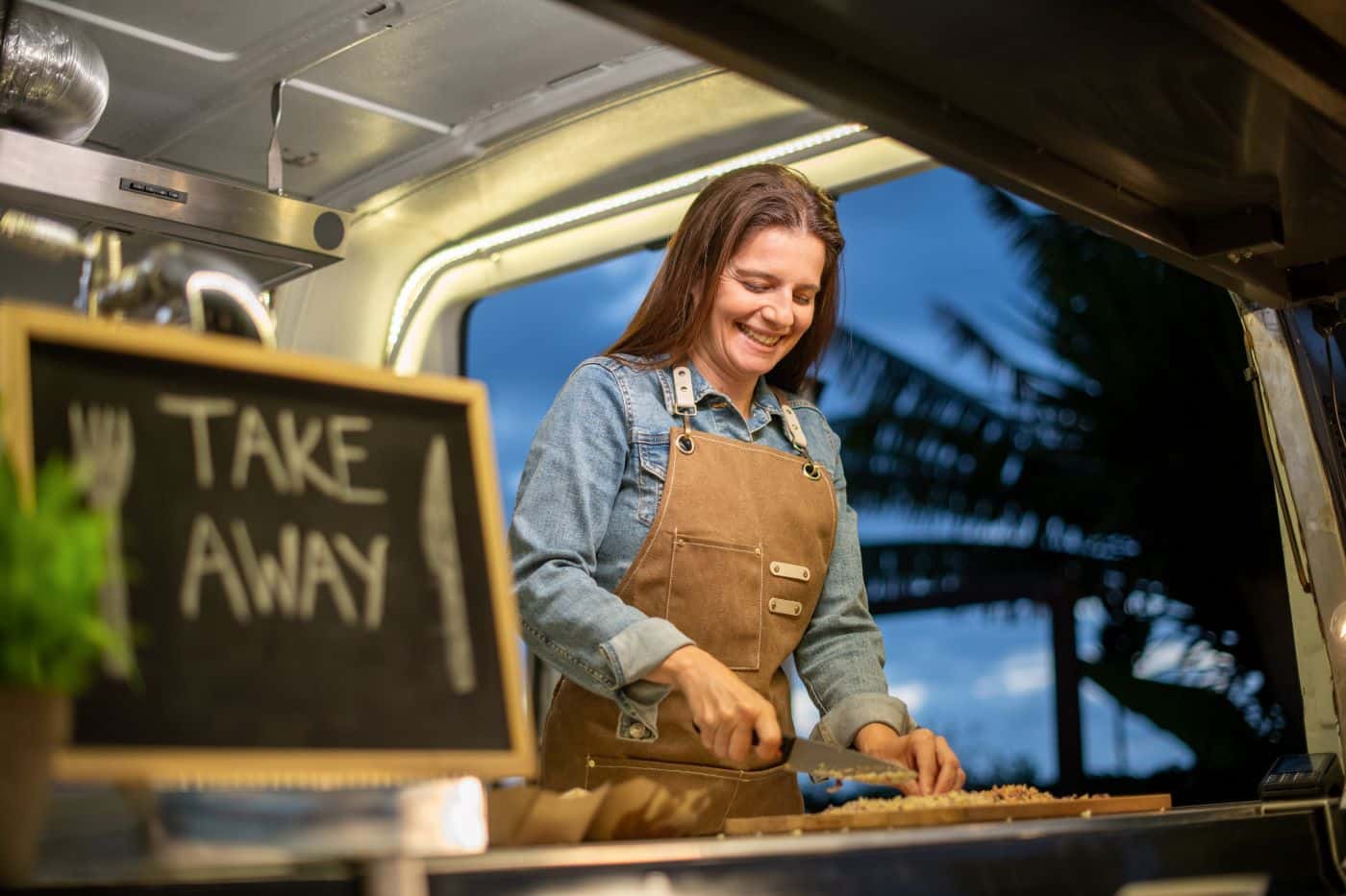 Myyjä valmistaa ruokaa ruokarekan luukulla Take away -taulun vieressä.
